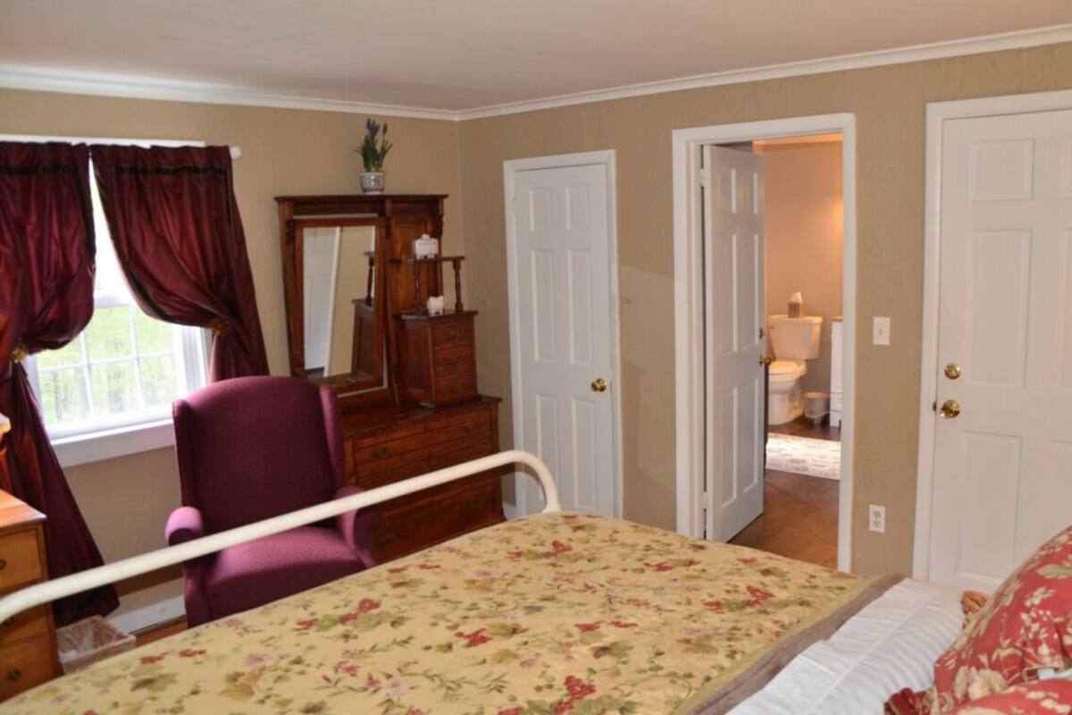 A bedroom with a bed, a chair, a dresser, and a mirror. There are two doors, one leading to a bathroom.