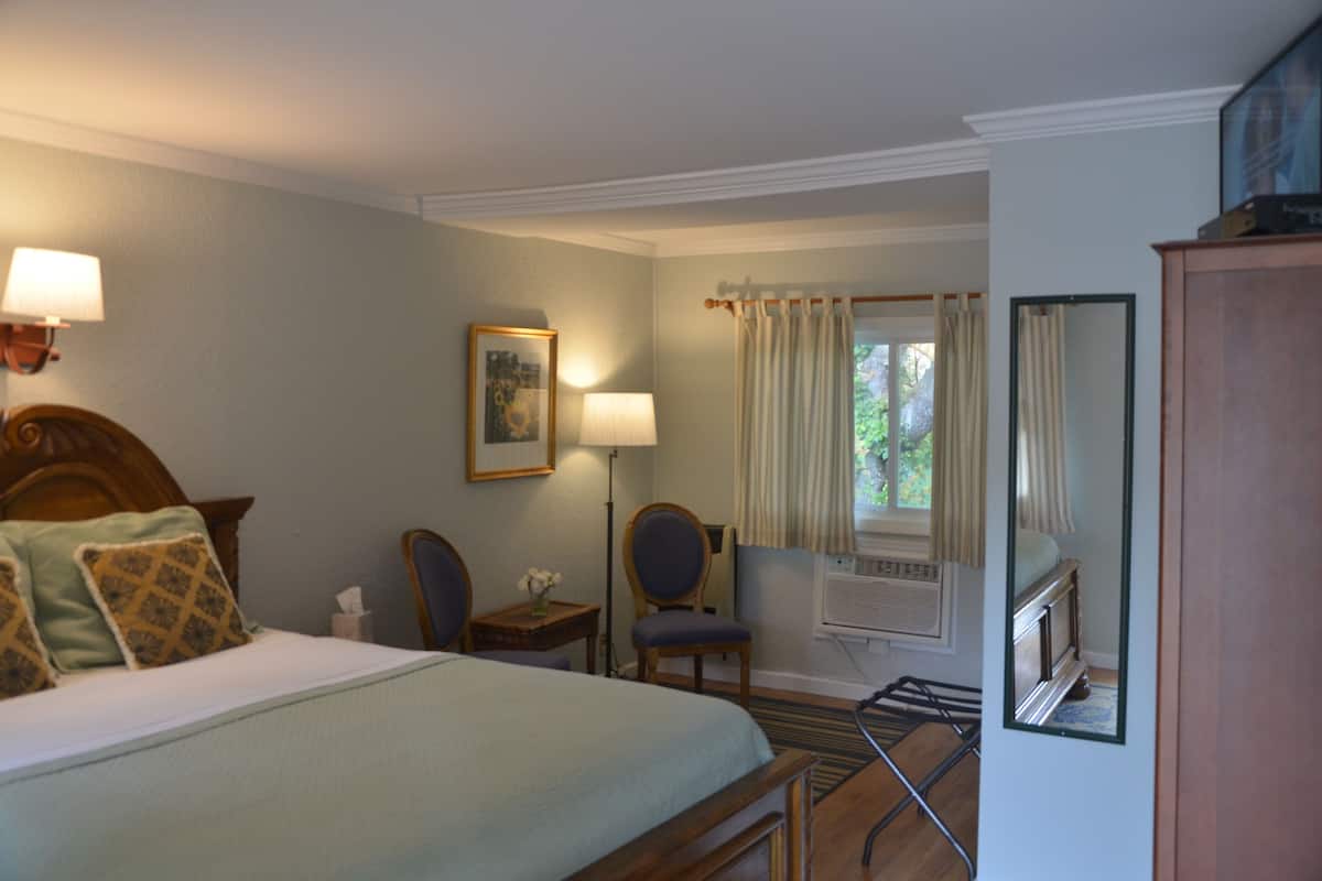 A hotel room with a bed, bedside lamps, a chair, a desk, a mirror, and a window with curtains.