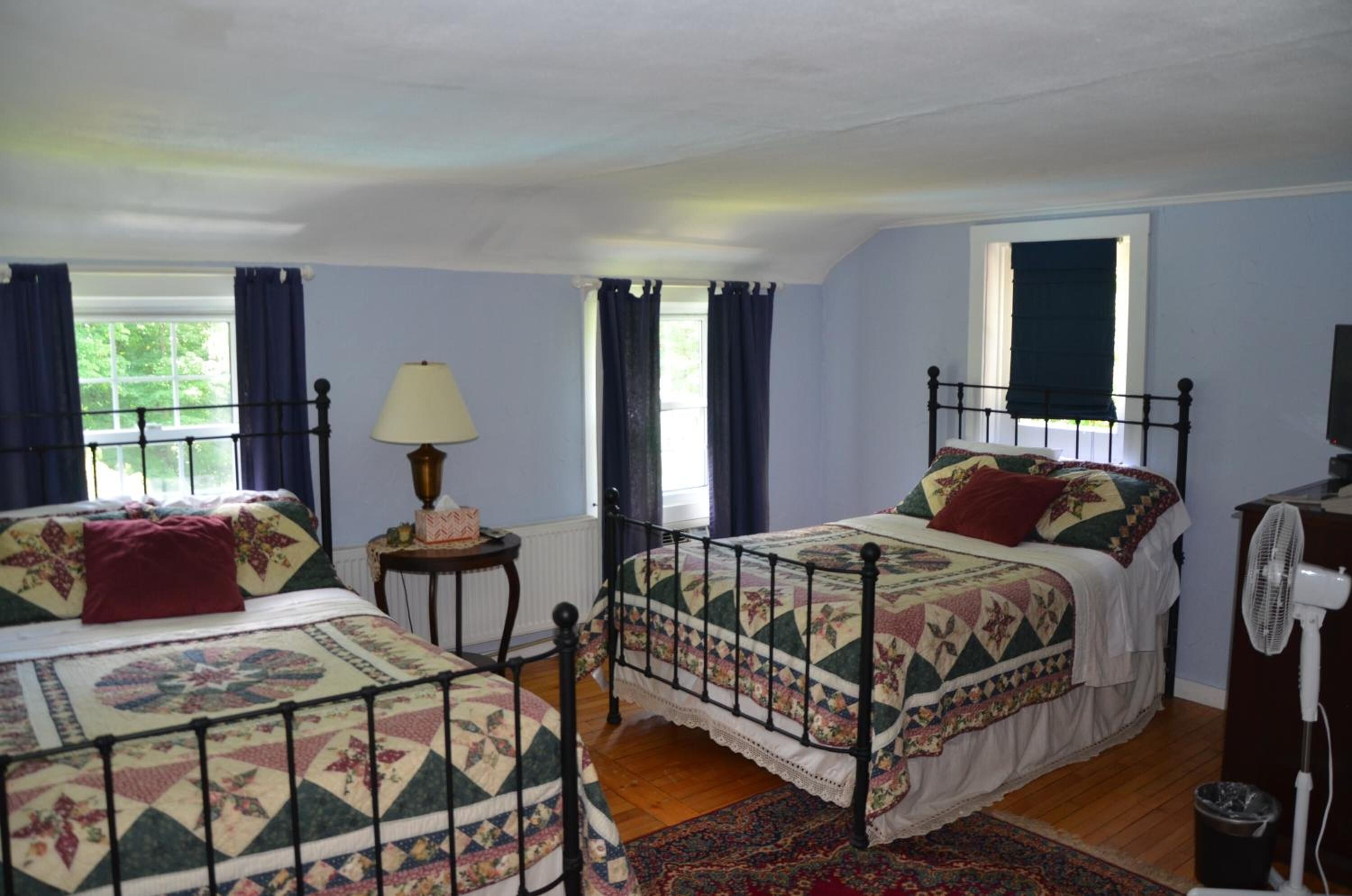 A bedroom with two single beds, a bedside table with a lamp, a window with curtains, a radiator, and a fan.