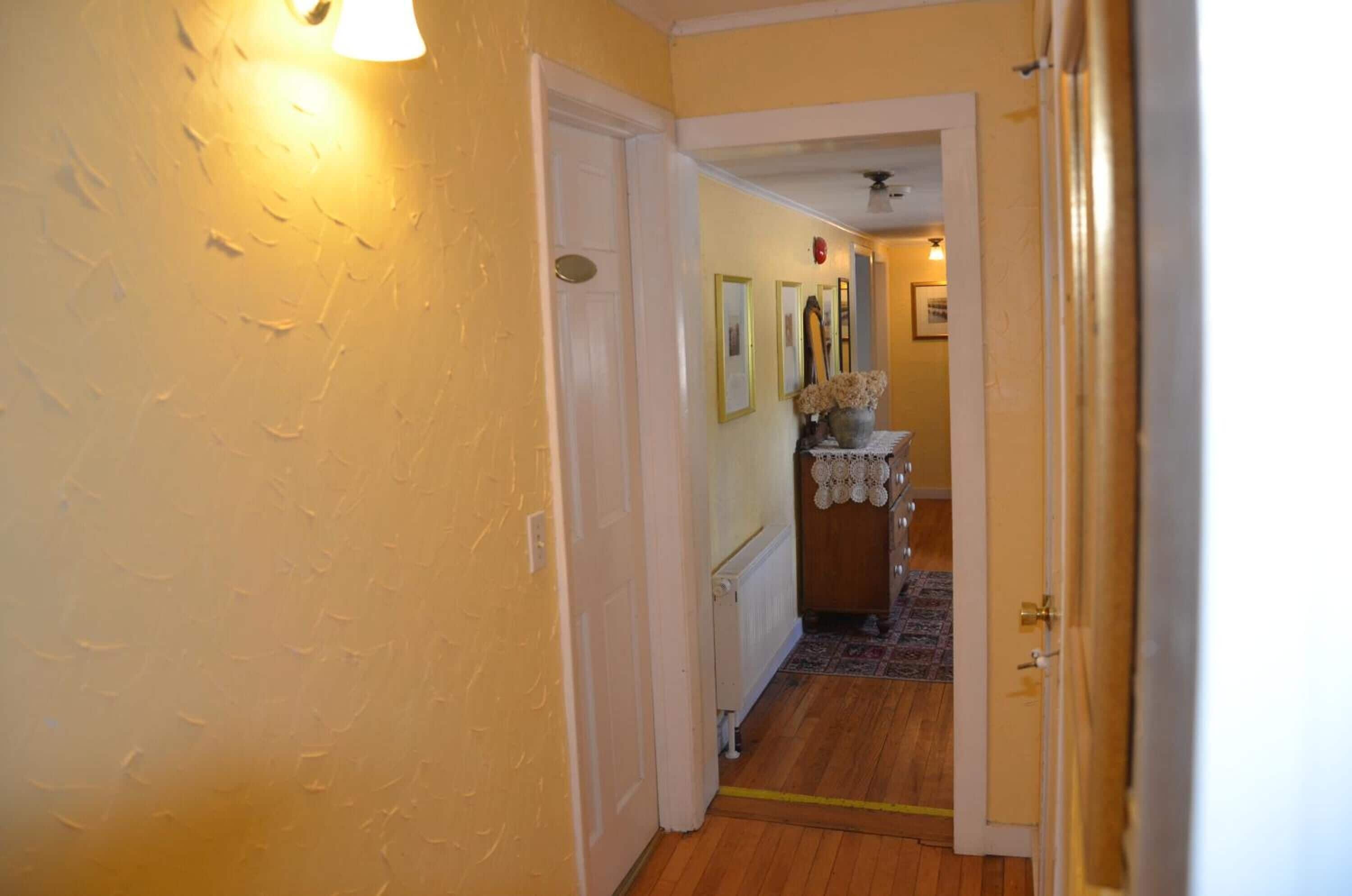 A hallway with a wooden floor, a door on the left, and a small table with a vase and framed pictures in the center.