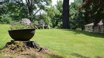 A grassy outdoor area with a large planter, two Adirondack chairs, and a house in the background.