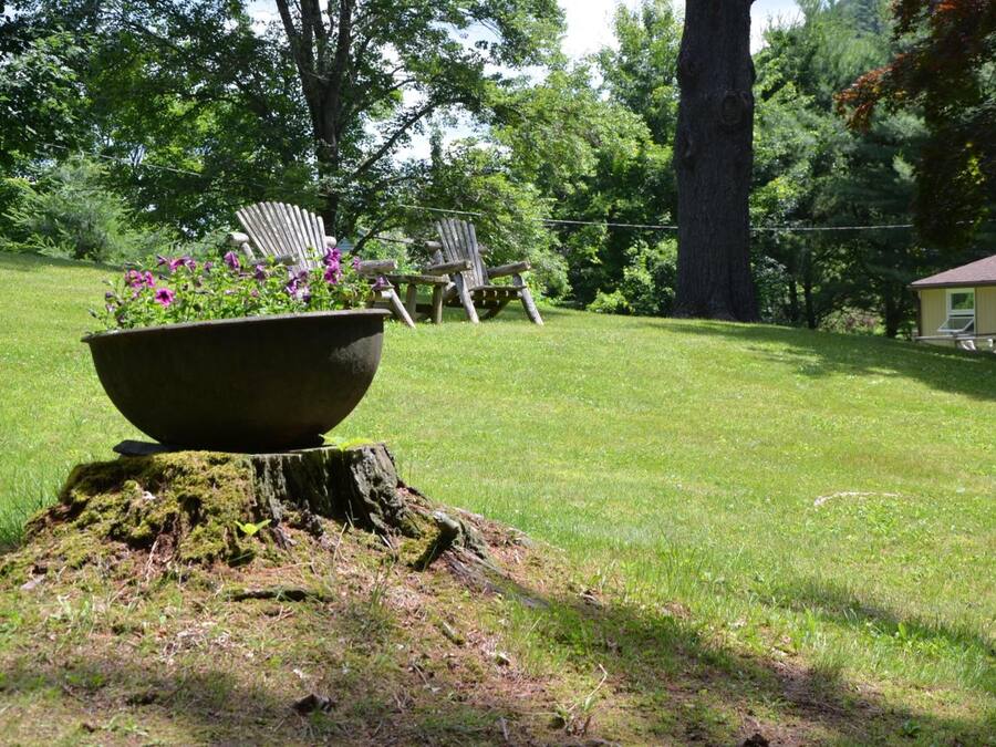 A grassy outdoor area with a large planter, two Adirondack chairs, and a house in the background.
