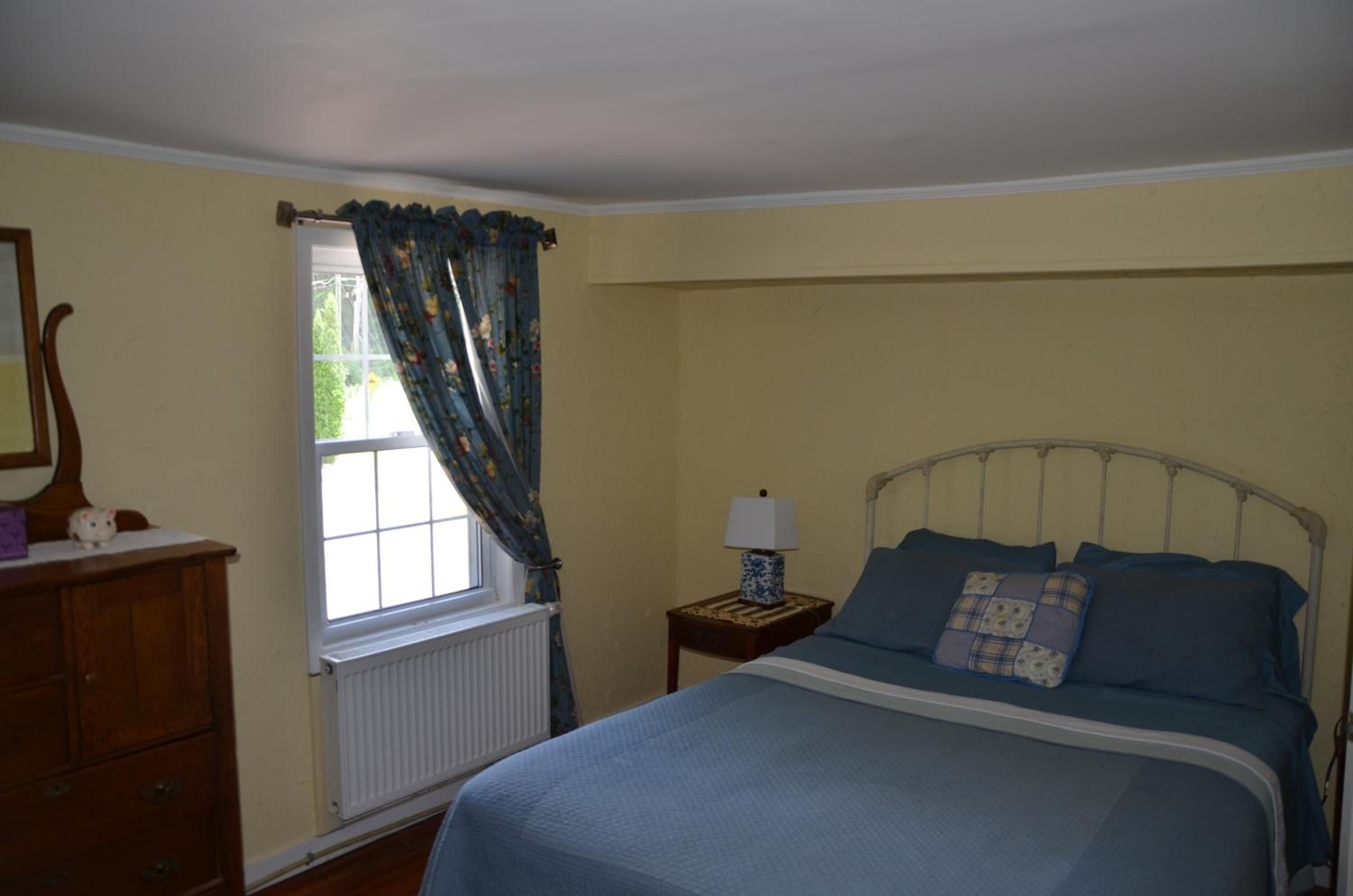 A bedroom with a bed, a dresser, a window with curtains, a nightstand with a lamp, and a radiator.