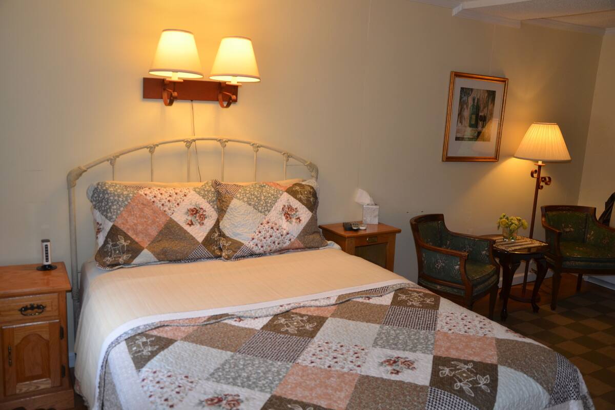 A neatly made bed with a quilt, two bedside lamps, a bedside table with a lamp, a chair, and a framed picture on the wall.