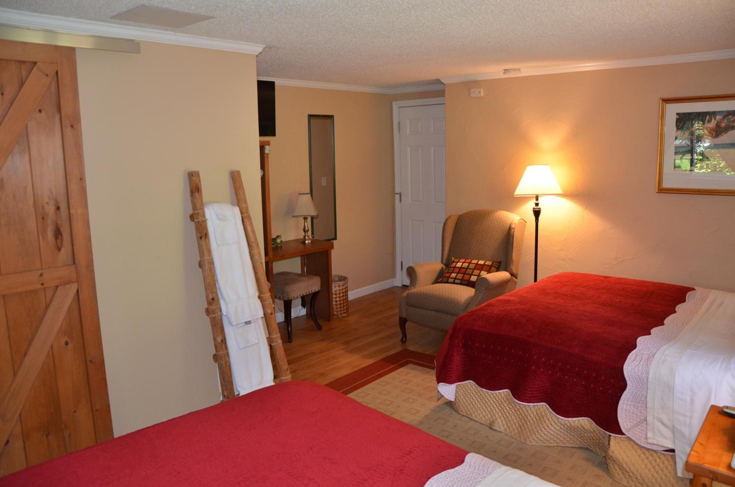 A bedroom with two beds, a chair, a desk, a lamp, and a door.