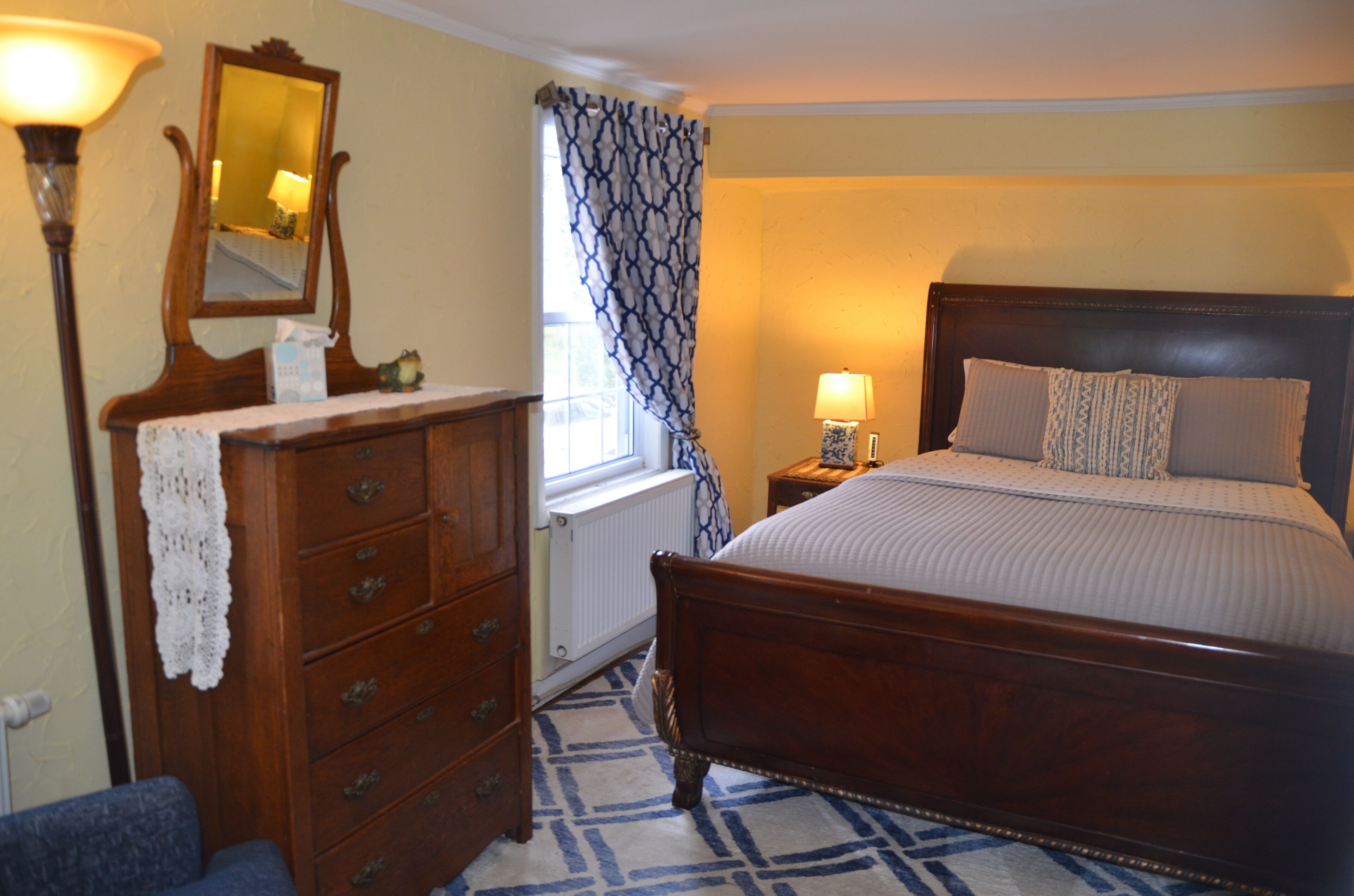 A bedroom with a wooden bed, a dresser, a mirror, a window with patterned curtains, and a lamp.
