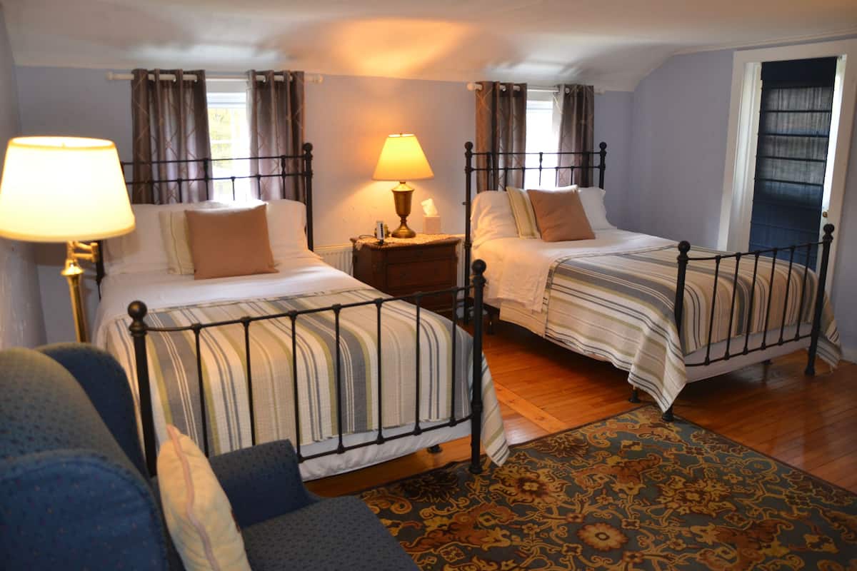 A room with two beds, a nightstand with a lamp, a patterned rug, and a window with curtains.