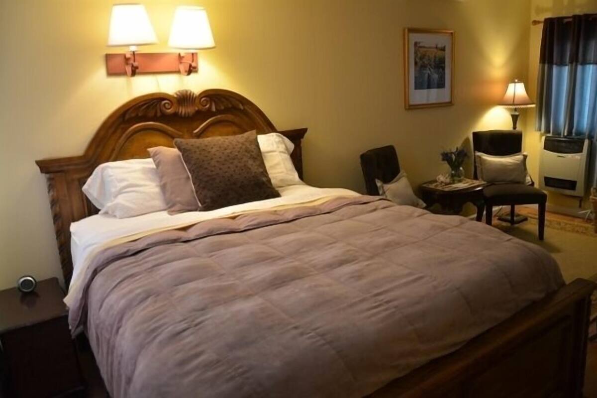 A neatly made bed with a wooden headboard, two bedside lamps, a chair, and a small table with a vase.