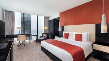 A hotel room with a large bed, a desk, a chair, and a view of a cityscape through large windows.