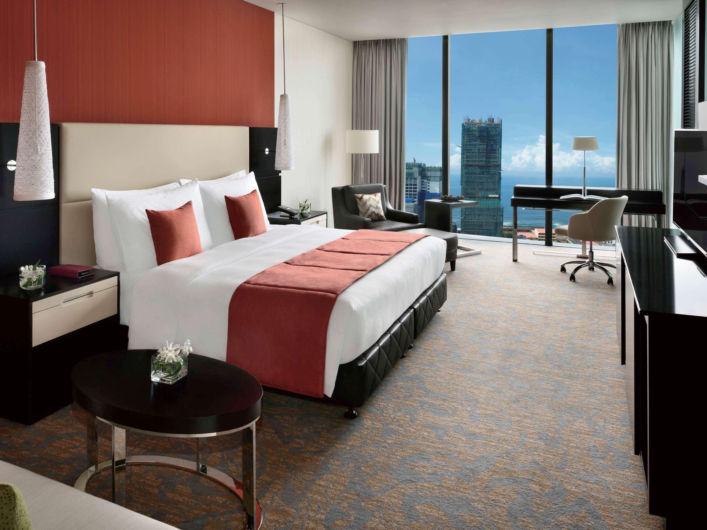 A hotel room with a large bed, a desk, a chair, a small table, and a view of a cityscape.