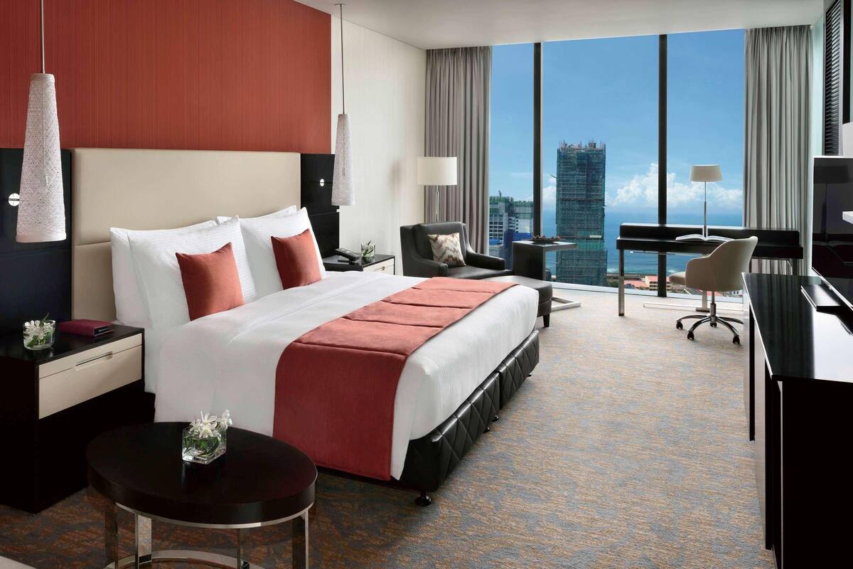 A hotel room with a large bed, a desk, a chair, a small table, and a view of a cityscape.