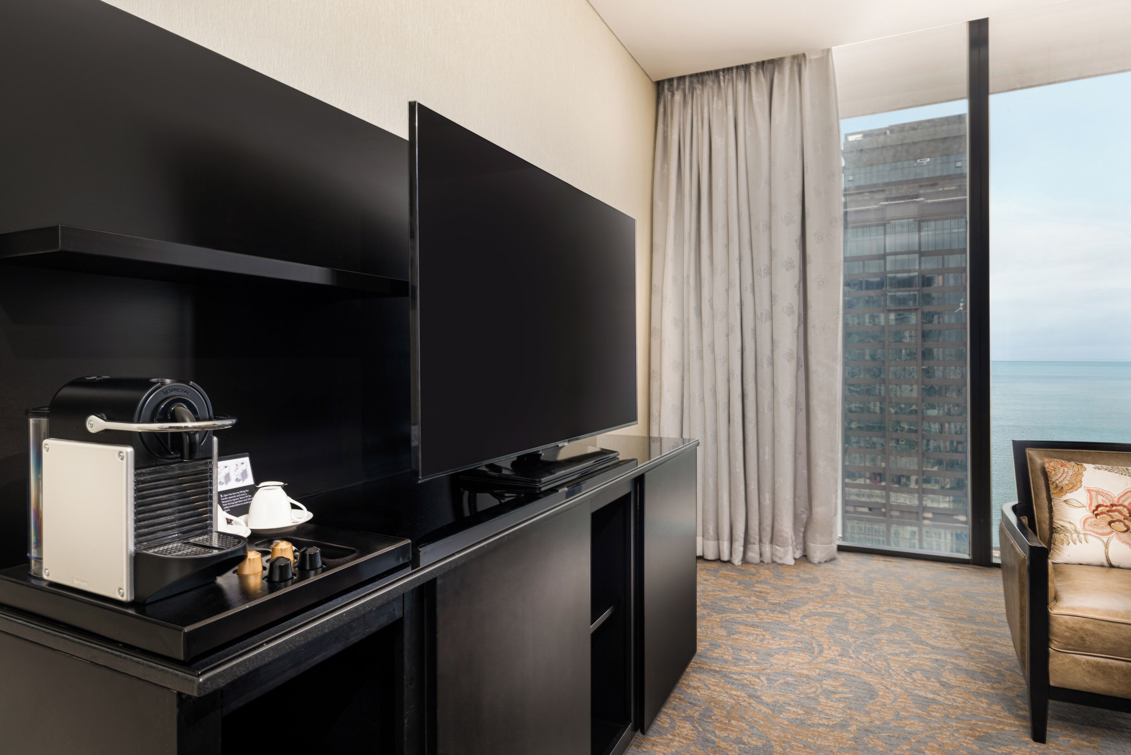 A hotel room with a flat-screen TV, a coffee machine, and a view of a building through a window.