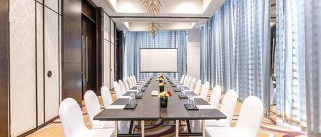 Un salón de conferencias con una mesa larga, sillas blancas, una pantalla de proyector y una alfombra estampada.