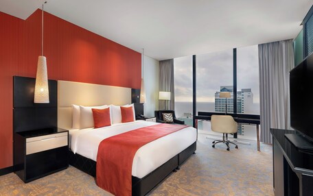 A hotel room with a large bed, a desk, a chair, a TV, and a view of the cityscape.