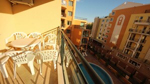 A balcony with white plastic chairs and a table, overlooking a swimming pool and adjacent buildings. - Menada Amadeus 3 Apartments (Sunny Beach)