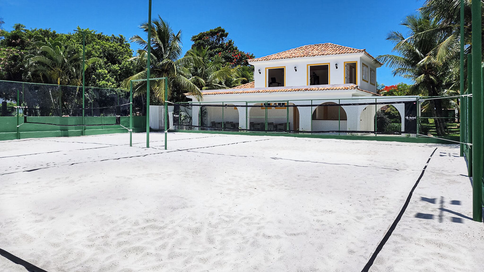A tennis court with a net, surrounded by a green fence. There is a building with a tiled roof and a white facade in the background.