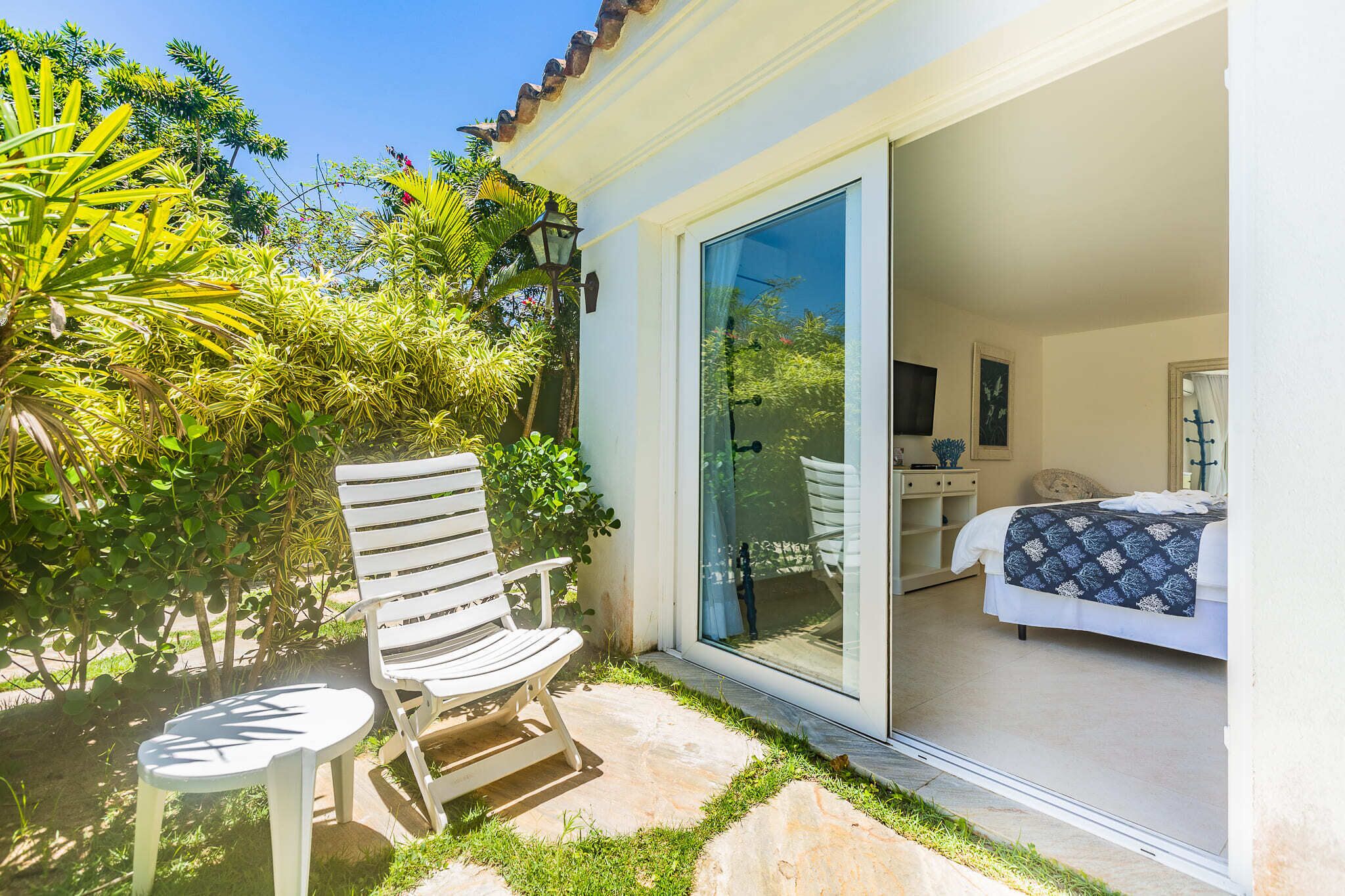 A patio with a white chair and table, a bed with a patterned blanket, and a view of greenery through a glass door.