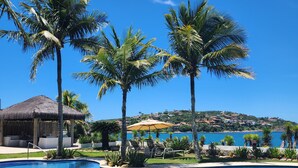 A swimming pool with palm trees, a thatched-roof hut, and a view of a coastal town. - Villa Raphael (Búzios)