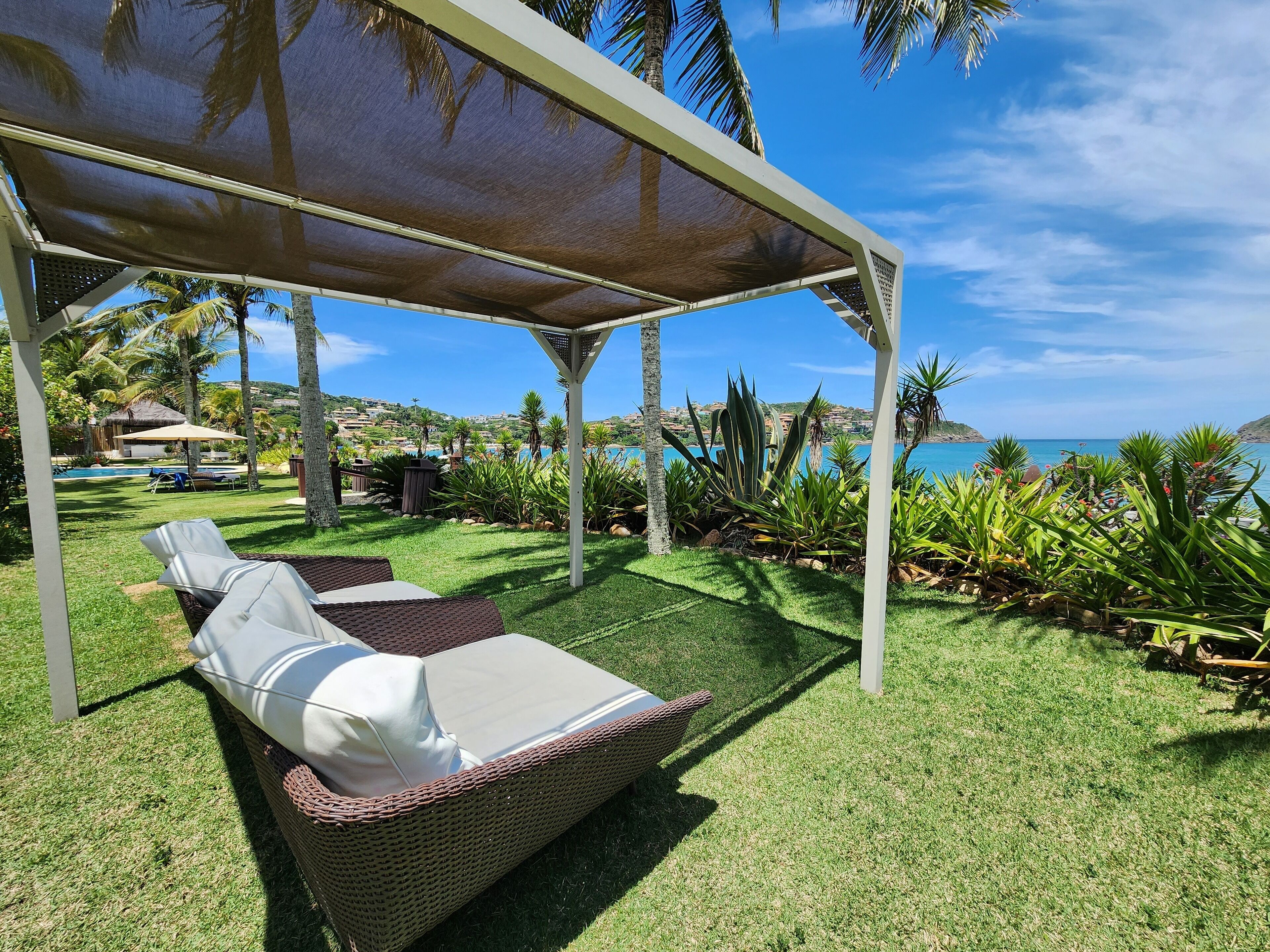 Un espace extérieur aménagé, abrité et doté de mobilier de jardin, comprenant un fauteuil de relaxation en osier et une table, le tout entouré d’une végétation luxuriante et offrant une vue sur la mer.