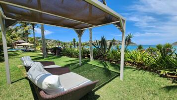A covered outdoor seating area with a wicker lounge chair and a table, surrounded by lush greenery and a view of the sea.