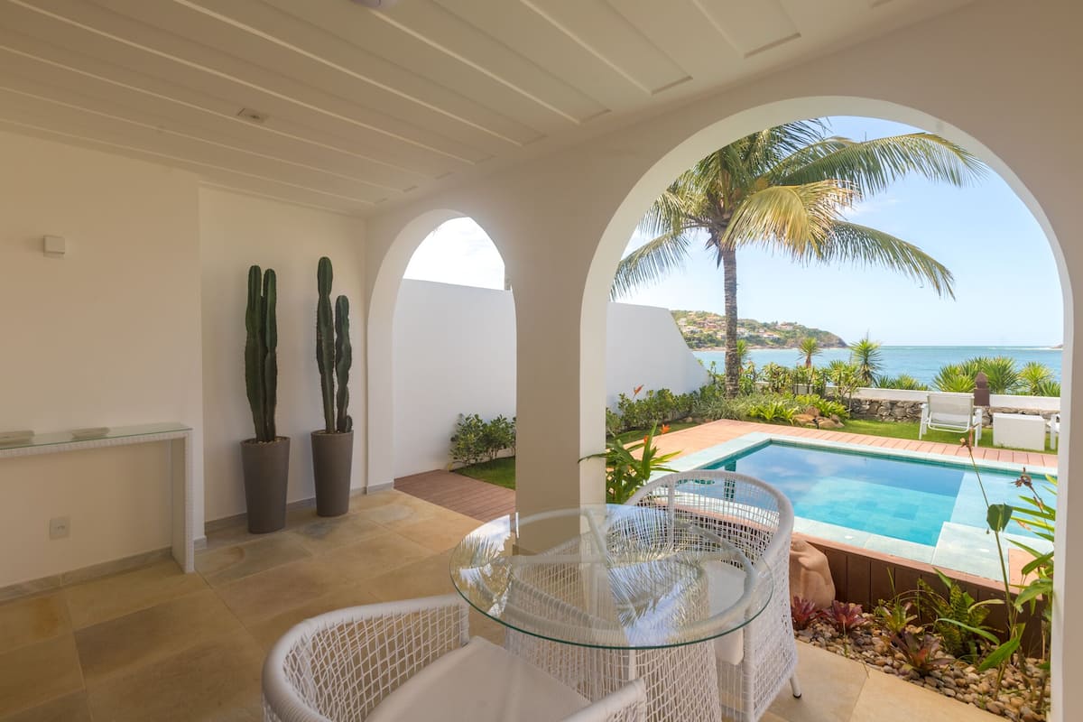 Un espace intérieur moderne, doté d'une table en verre et de chaises en osier blanc, qui donne accès à une zone de piscine offrant une vue sur la mer et les palmiers.