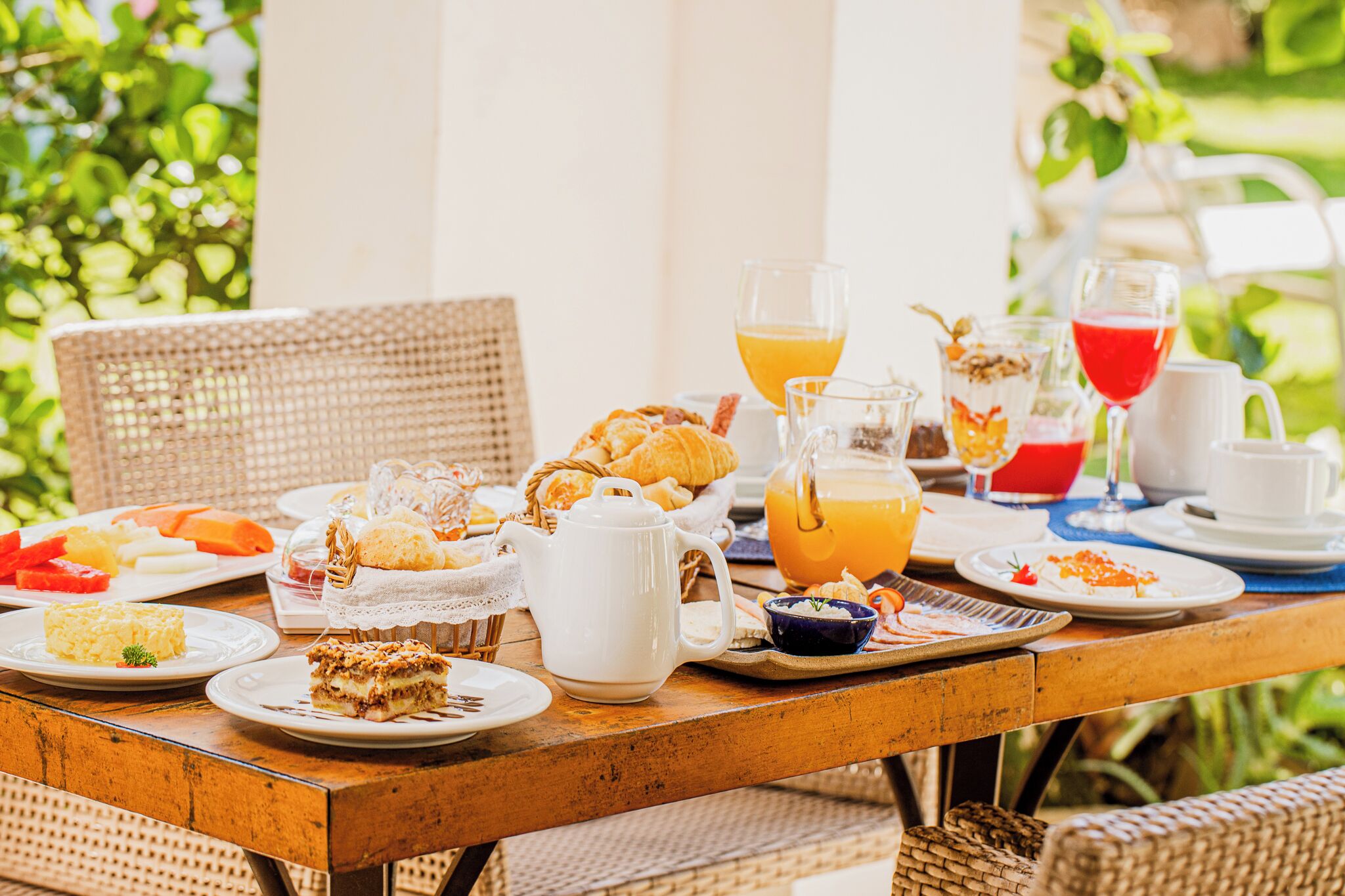 Una mesa de madera dispuesta con alimentos para el desayuno, como pasteles, fruta y bebidas, rodeada de sillas de mimbre.