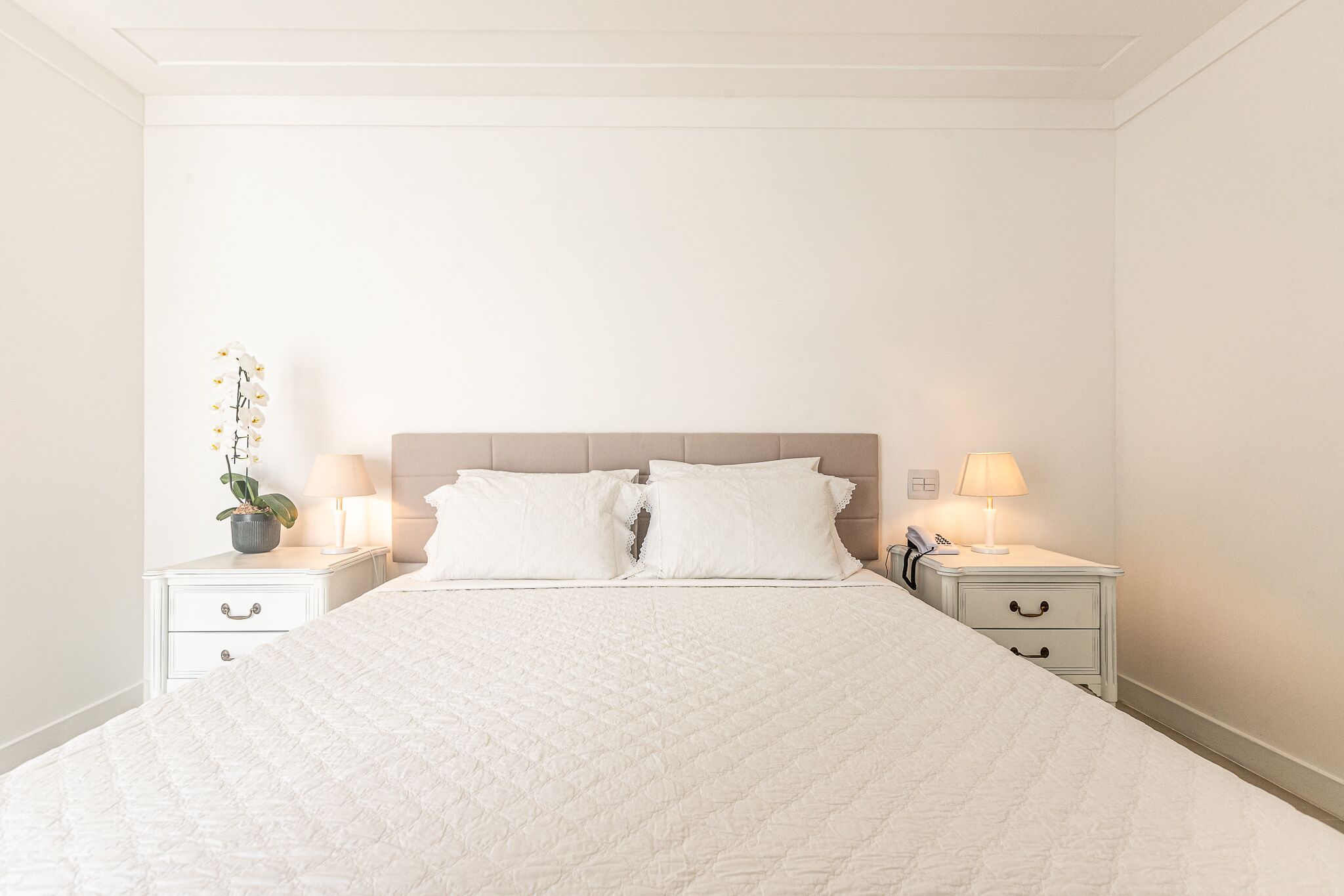 A neatly made bed with white bedding, two bedside tables with lamps, and a potted plant on one table.
