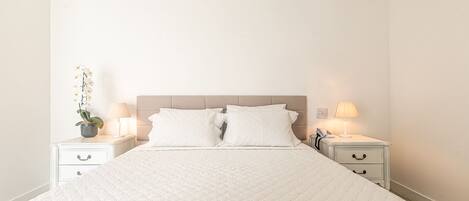 A neatly made bed with white bedding, two bedside tables with lamps, and a potted plant on one table.
