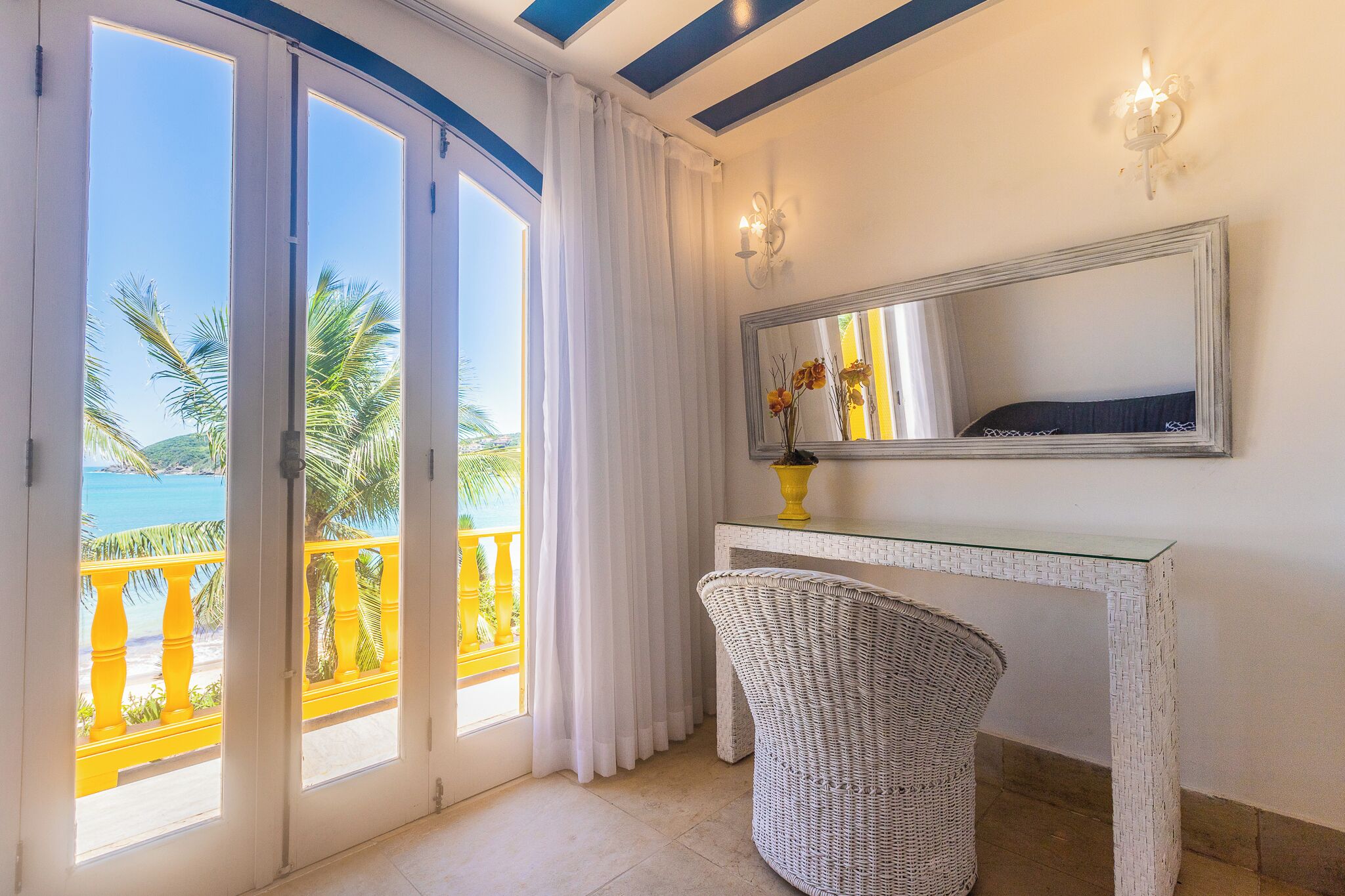 A room with a glass door leading to a balcony, a wicker chair, a white console table, and a mirror on the wall.