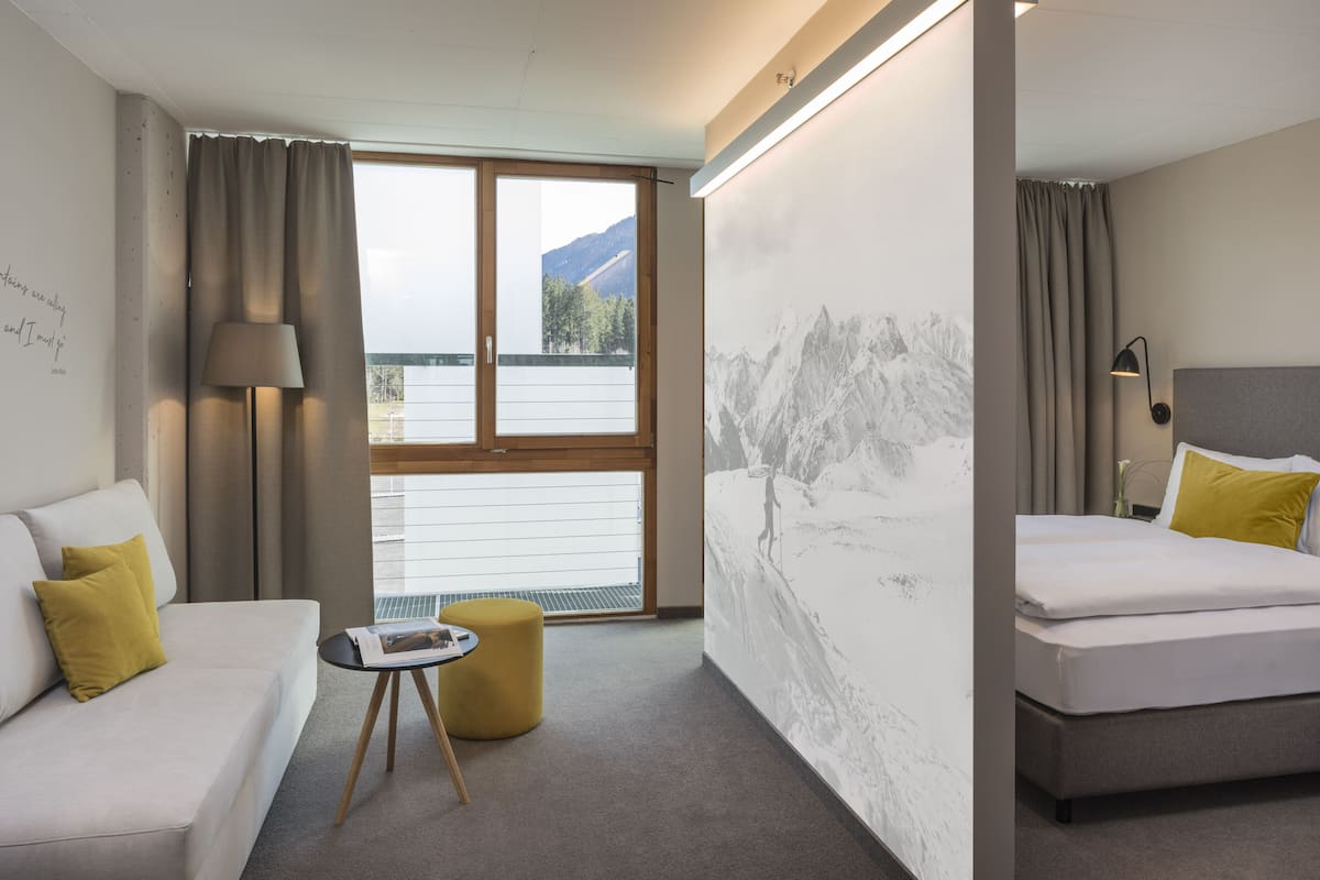 Habitación de hotel con sofá, cama, una mesita redonda, lámpara y vistas a montañas a través de un ventanal grande.