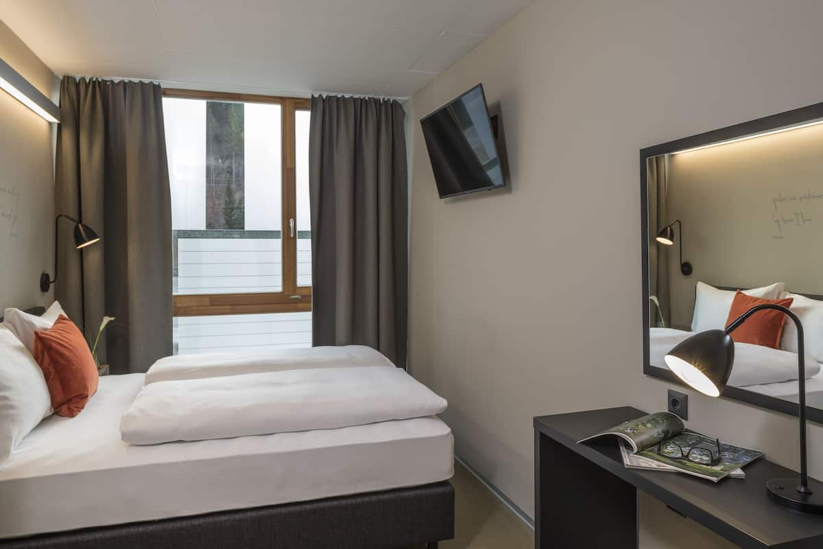 Habitación de hotel con cama, escritorio con lámpara, televisor de pantalla plana y ventana con cortinas.