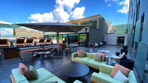 Ein moderner Außenbereich mit Sitzgelegenheiten, darunter Sofas, Stühle und ein runder Tisch, ausgestattet mit einem großen Sonnenschirm und Glasüren zum Pool.