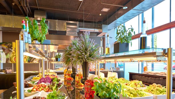 Ein Buffet mit verschiedenen Speisen, darunter Salate, Obst und Gemüse, in einem gut beleuchteten Innenraum mit großen Fenstern und moderner Küchenausstattung im Hintergrund.