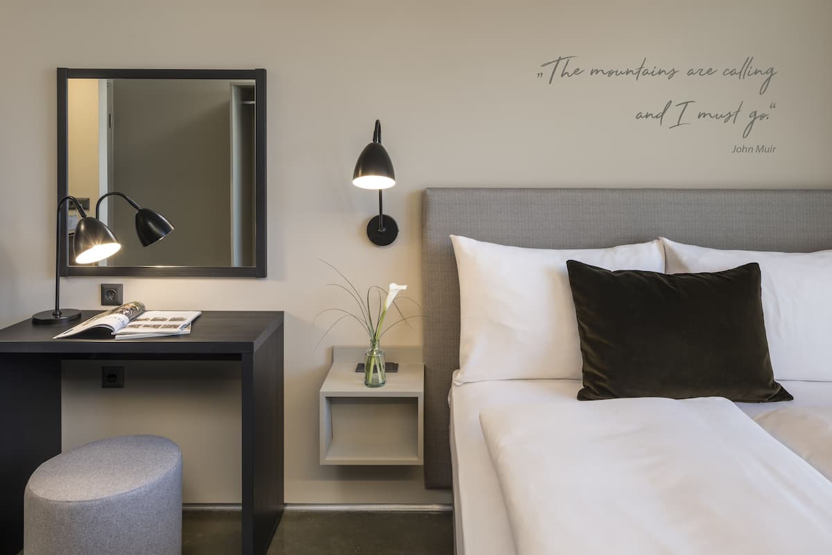 Habitación de hotel ordenada con cama, mesita de noche, lámpara y una frase decorativa en la pared.
