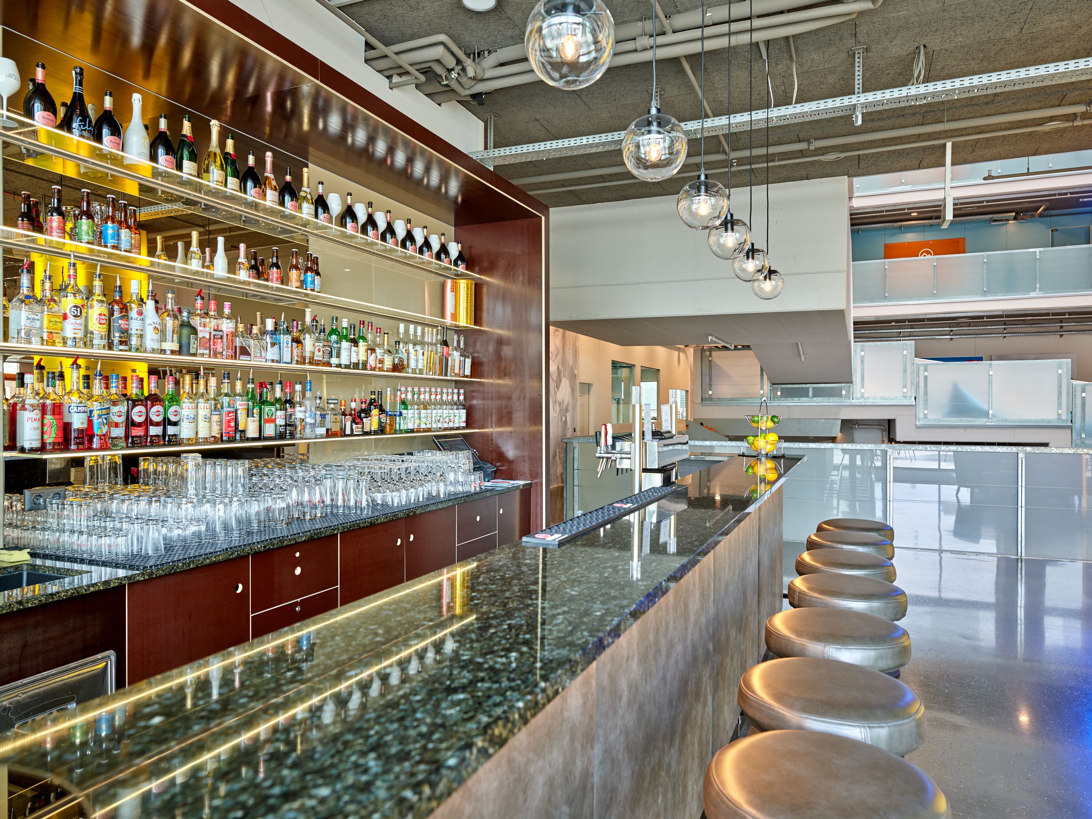 A bar area with shelves of liquor bottles and glasses, a marble countertop, and bar stools.