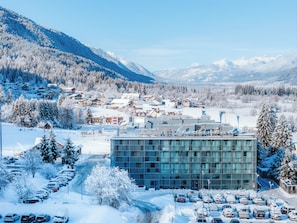 A hotel with a modern glass facade, surrounded by snow-covered trees and mountains. - Arena Franz Ferdinand Nassfeld (Hermagor-Pressegger See)