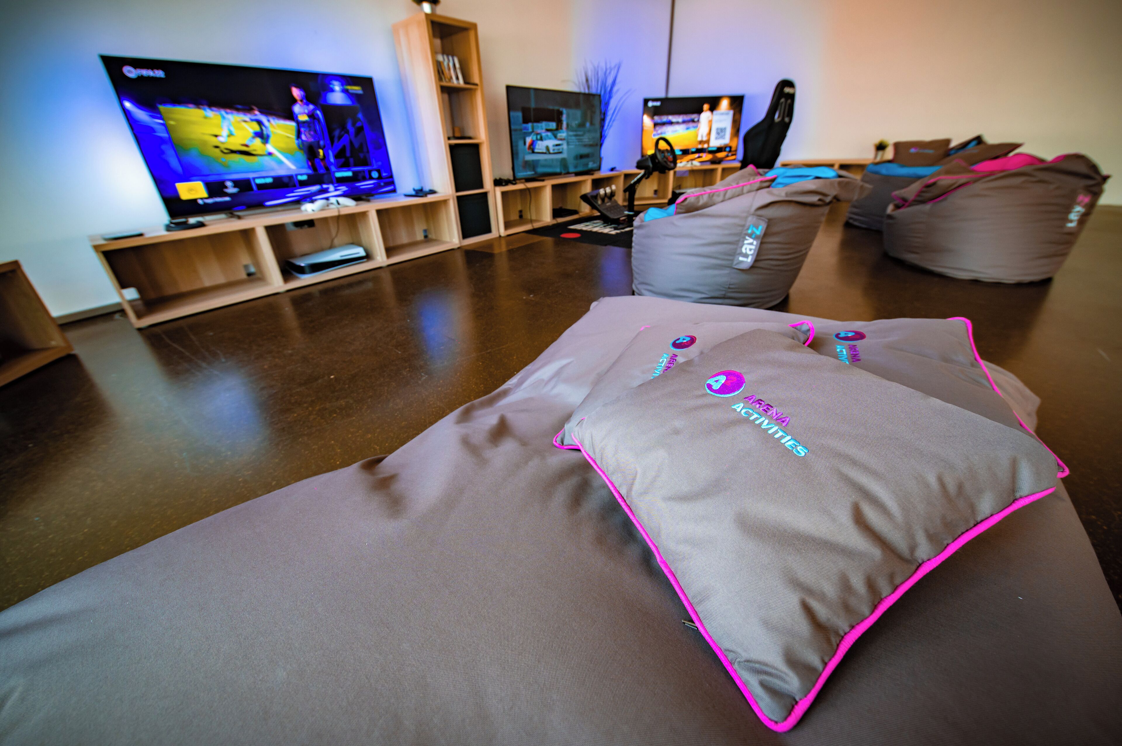 Ein Zimmer mit zwei großen Flachbildfernsehern auf Holzständer, ein Sofa mit rosa und grauem Überwurf und mehreren Sitzsäcken.