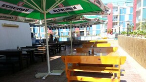 Outdoor seating with umbrellas, a brick floor, and a pool in the background. - Menada Elit IV Apartments (Sunny Beach)