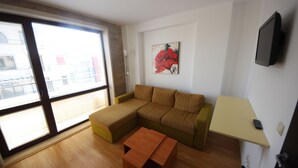 A living room with a sectional sofa, a small table, a painting on the wall, and a TV mounted on the wall. - Menada Amara Apartments (Sunny Beach)