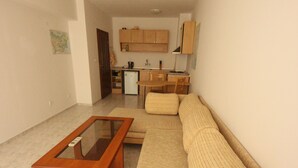 A living room with a glass-top coffee table, a beige sofa, and a kitchen area in the background.