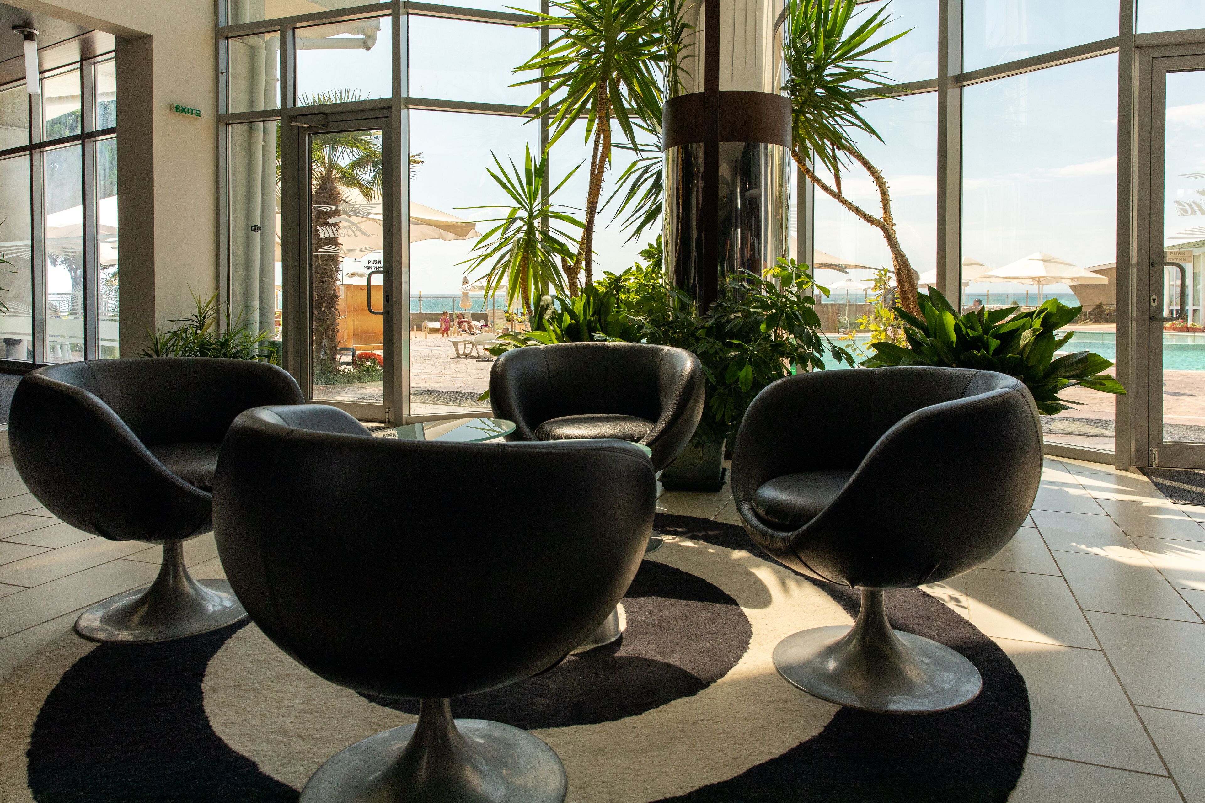 Un hall d’entrée moderne avec des fauteuils en cuir noir, une grande porte vitrée donnant accès à un espace piscine et des plantes d’intérieur.