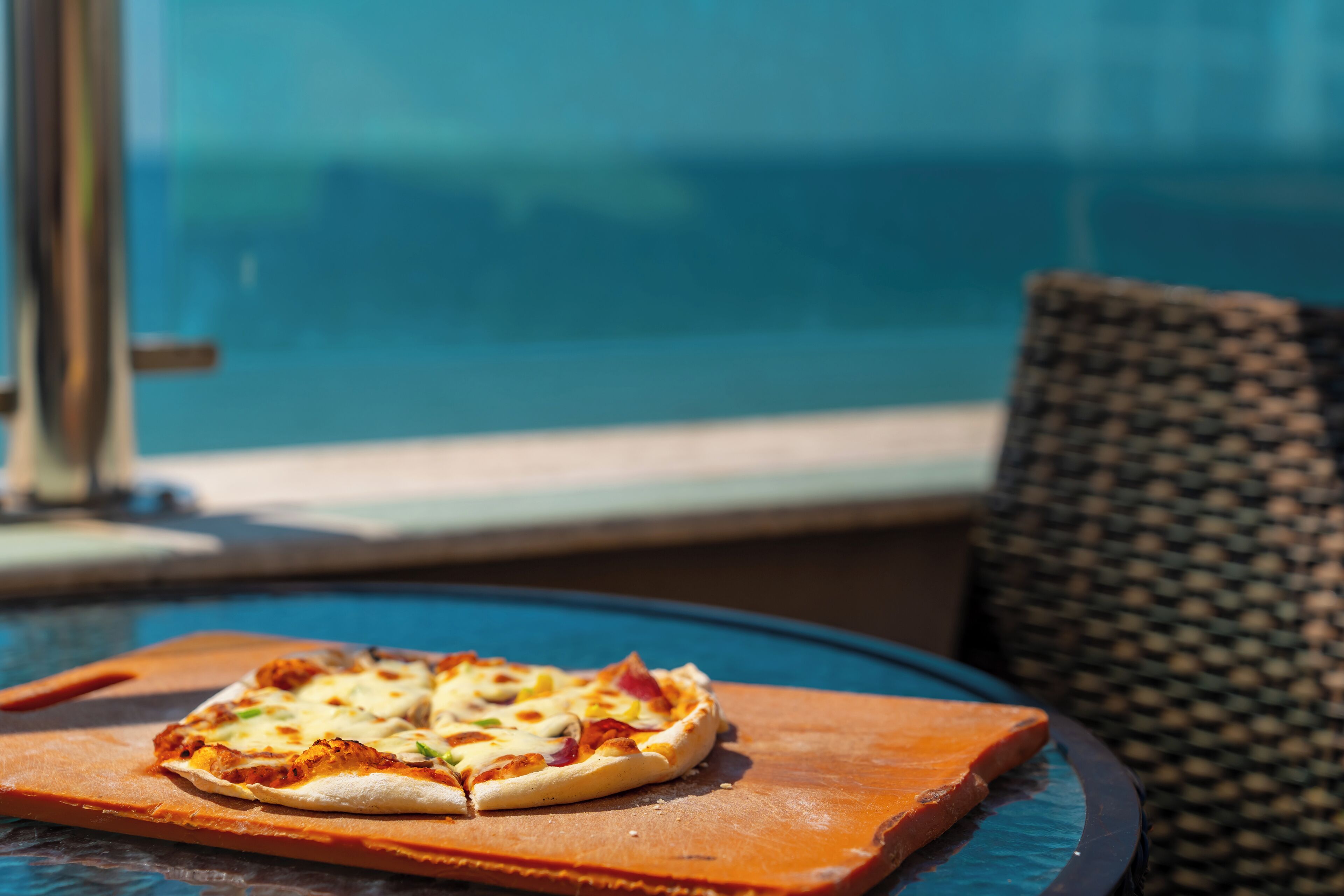 Una pizza sobre una tabla de madera, sobre una mesa de vidrio, junto a una ventana con vista a una piscina.
