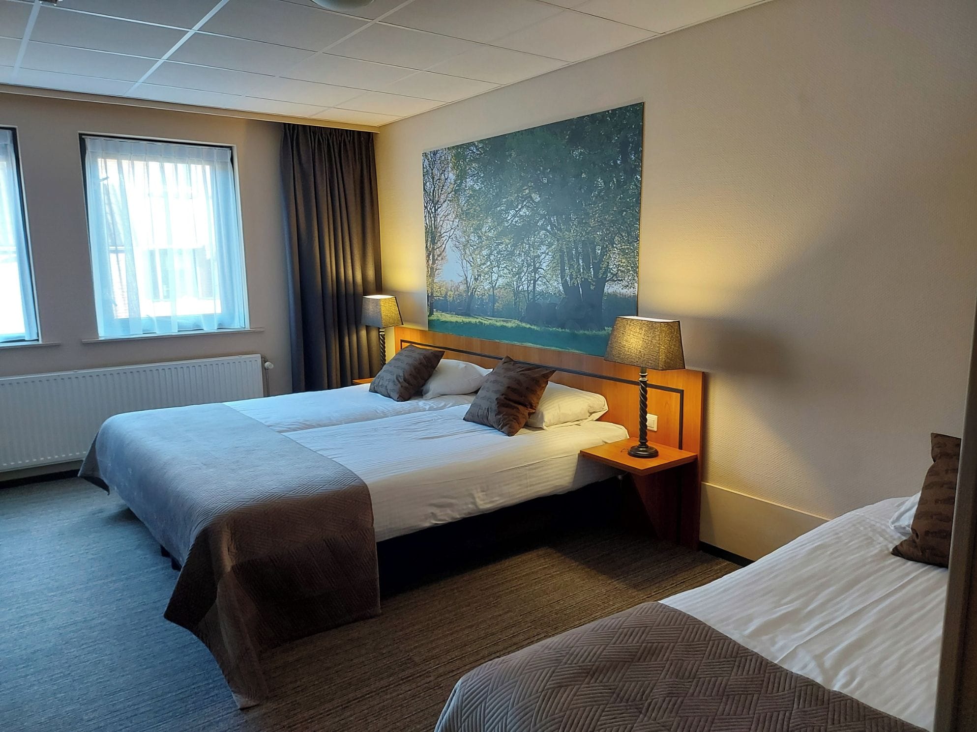 Habitación de hotel con cama doble, cama individual, un cuadro de un bosque, una ventana con cortinas y una lámpara en la mesita de noche.