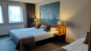 Habitación de hotel con cama doble, cama individual, un cuadro de un bosque, una ventana con cortinas y una lámpara en la mesita de noche.