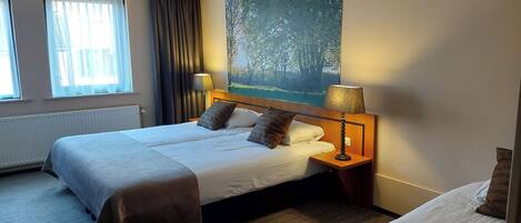 A hotel room with a double bed, a single bed, a painting of a forest, a window with curtains, and a lamp on the nightstand.