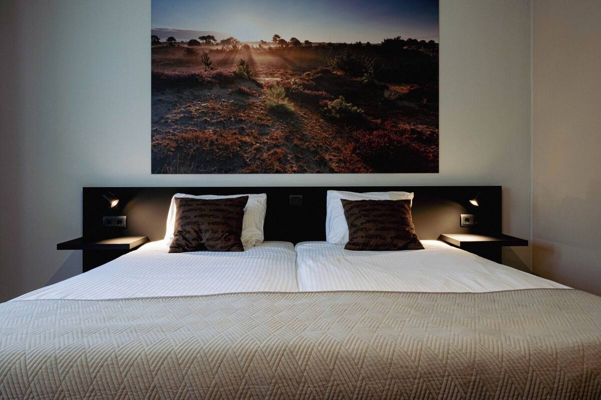 A neatly made bed with a patterned bedspread and two pillows, flanked by bedside tables with lamps, in a room with a large landscape painting on the wall.