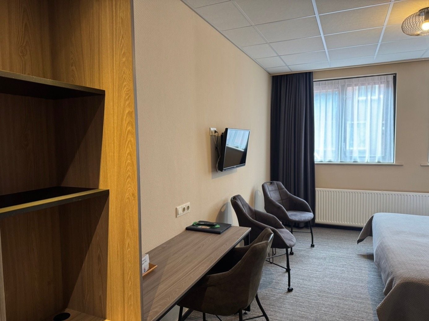 A hotel room with a wooden desk, two chairs, a bed, a TV, and a window with curtains.