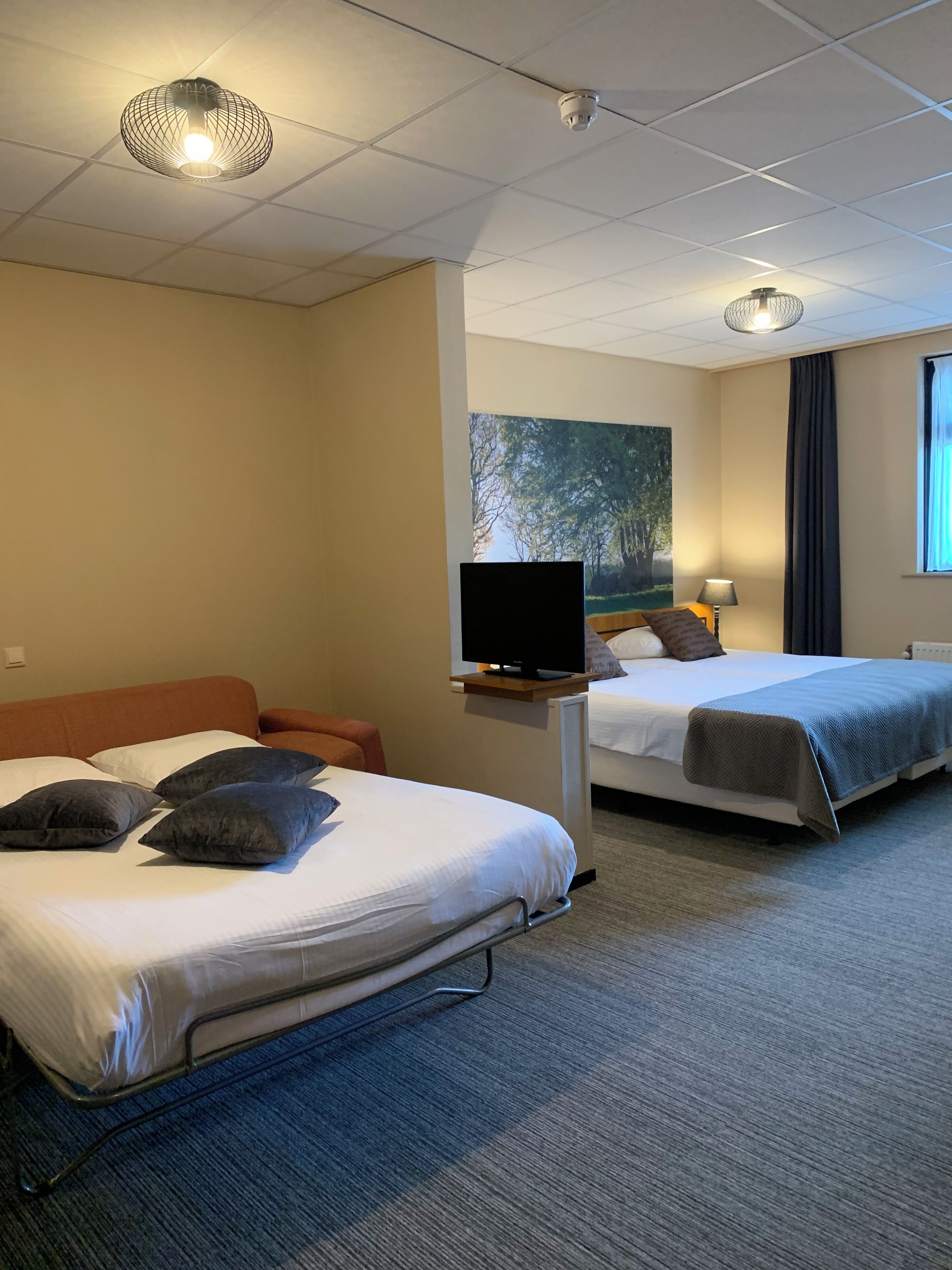 Ett hotellrum med två sängar, en tv på ett stativ, en tavla på väggen och ett fönster med gardiner.