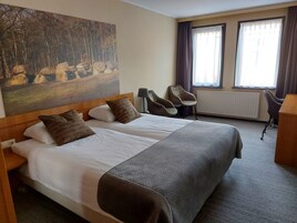 Habitación de hotel con dos camas, un cuadro de un paisaje forestal, un escritorio, dos sillas y una ventana con cortinas.