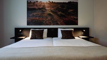 Een net opgemaakt bed met een beddekleed met dessin en twee kussens, met nachtkastjes met lampen ernaast, in een kamer met een groot landschap op de muur.